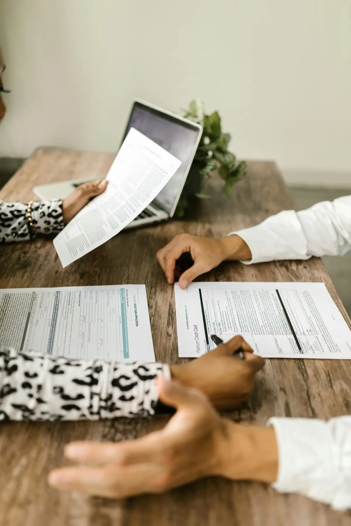 Deux personnes discutent de documents sur une table en bois, avec un ordinateur portable et des plantes en arrière-plan. Divers papiers sont éparpillés autour de la table.